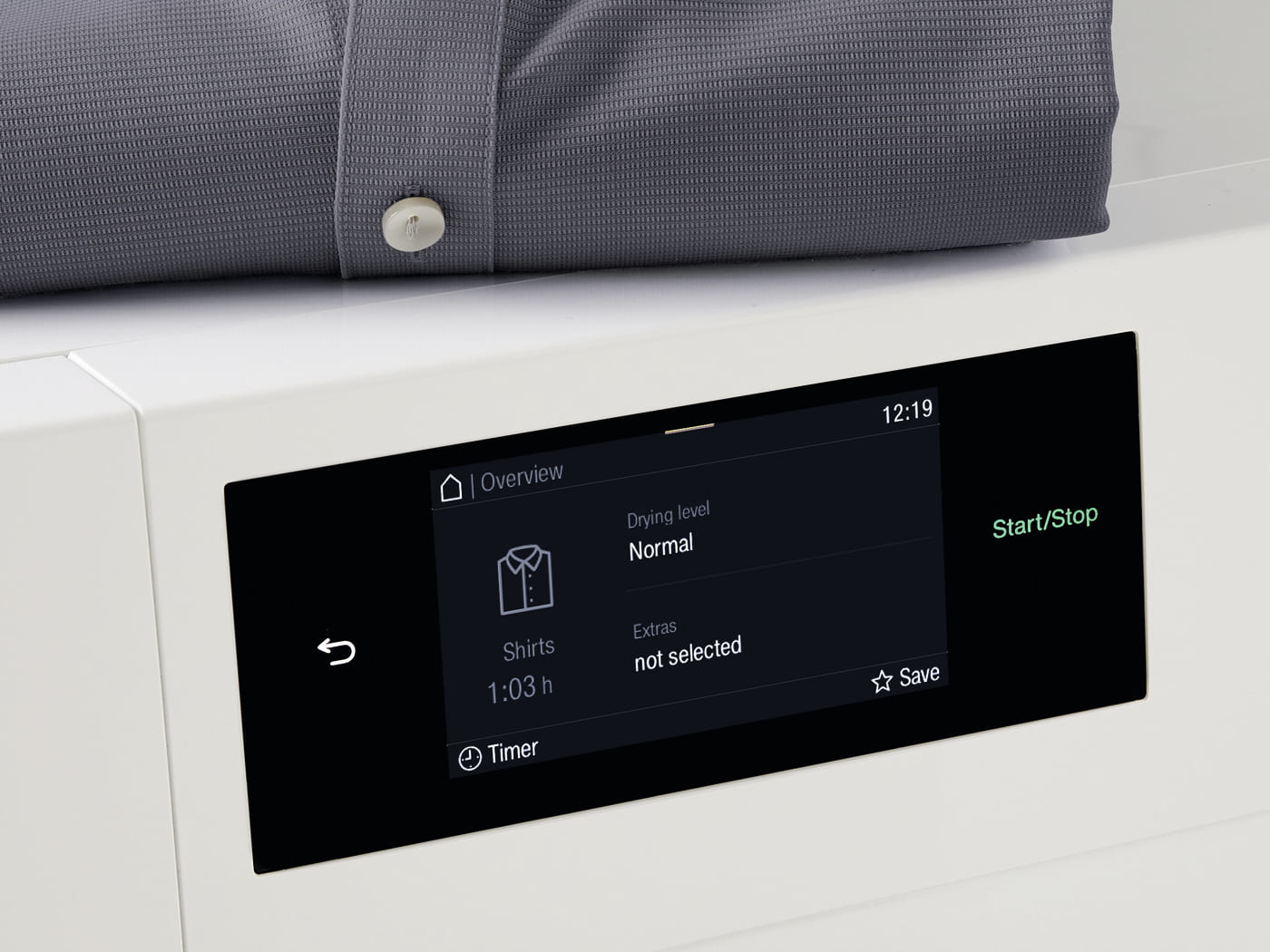 Close-up of a white appliance with a black touchscreen displaying 'Overview'—Drying level Normal, Shirts 1:03 h, Extras not selected; Start/Stop on the right.
