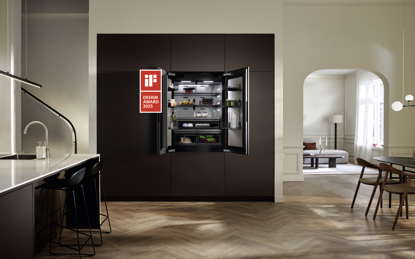 Open built-in dark kitchen cabinet with a refrigerator door swung open to reveal organized shelves; iF Design Award 2025 badge on the left door; dining area and living room beyond.