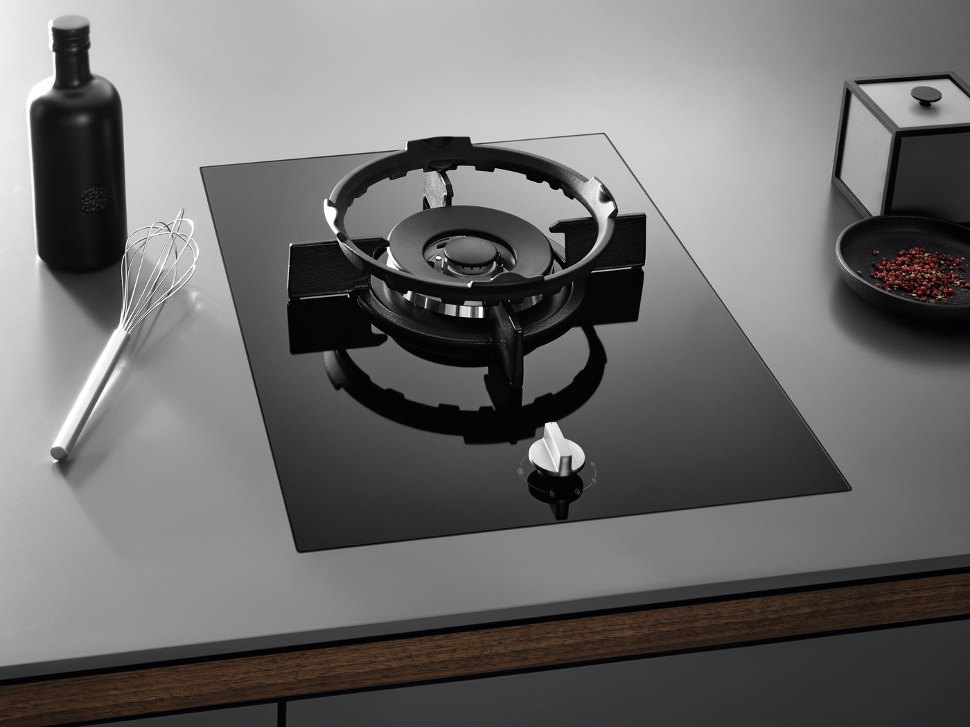 Glass black gas cooktop on a gray counter, with a whisk, a dark bottle, and a small dish of peppercorns nearby.