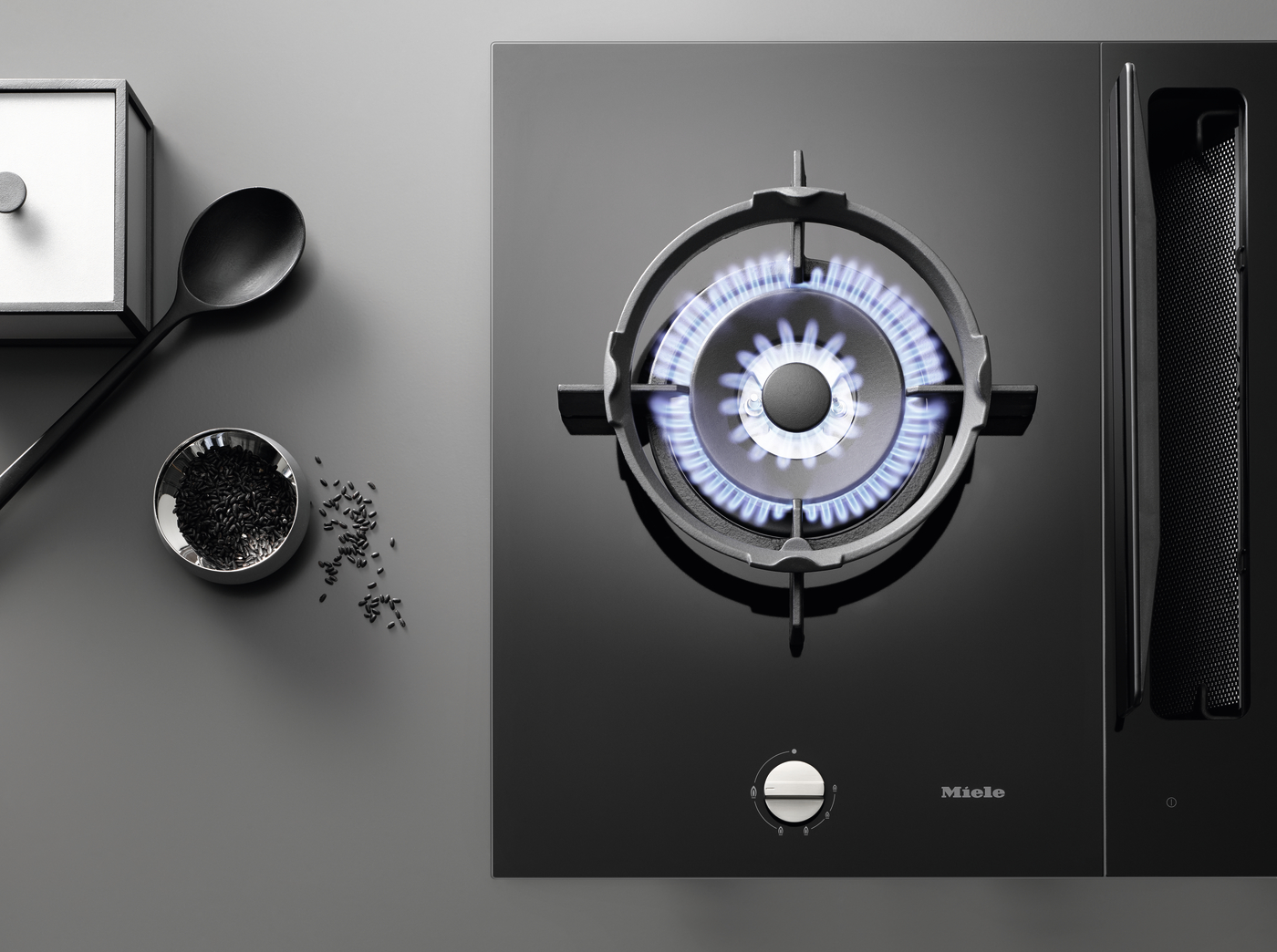 Top-down view of a black glass gas cooktop with a blue-flame burner; a spoon and a small bowl of black rice sit on the gray counter nearby (Miele logo visible).