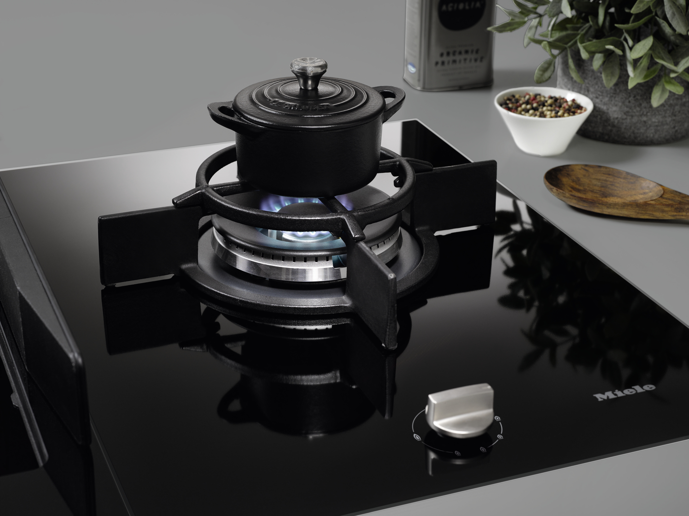 Gas stove with a pot simmering on the front burner; glossy black surface and nearby kitchen items (olive oil, peppercorns, wooden spoon) on the counter nearby.