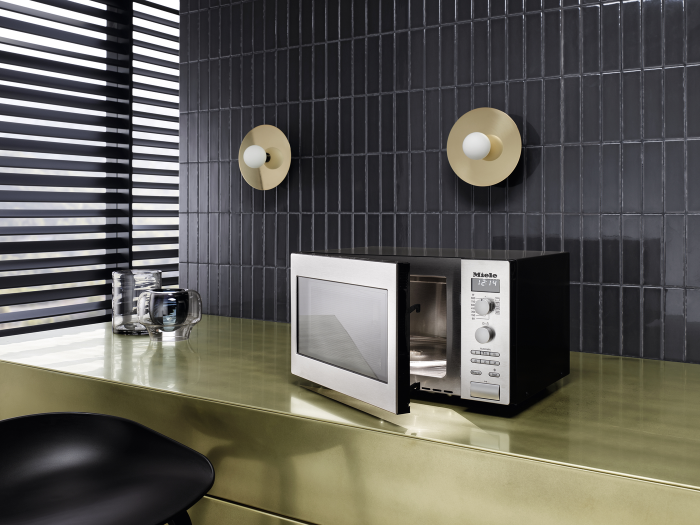 Sleek microwave on a glossy gold countertop in a modern kitchen with dark tiled wall and circular brass wall lights above.