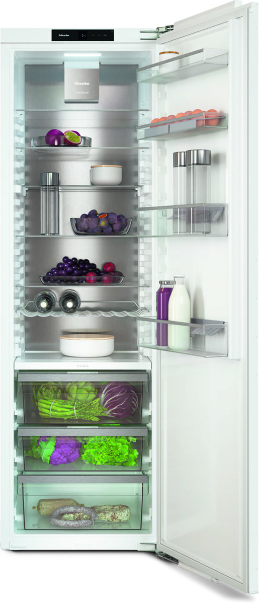 Open refrigerator door revealing multiple shelves and produce: grapes, peppers, leafy greens, milk, and jars arranged with clear drawers below.