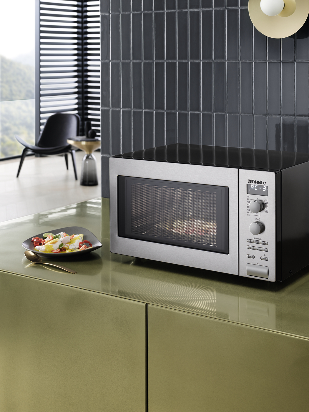 Stainless steel microwave on a glossy olive-green counter with a plate of colorful salad and a golden spoon nearby in a modern kitchen.