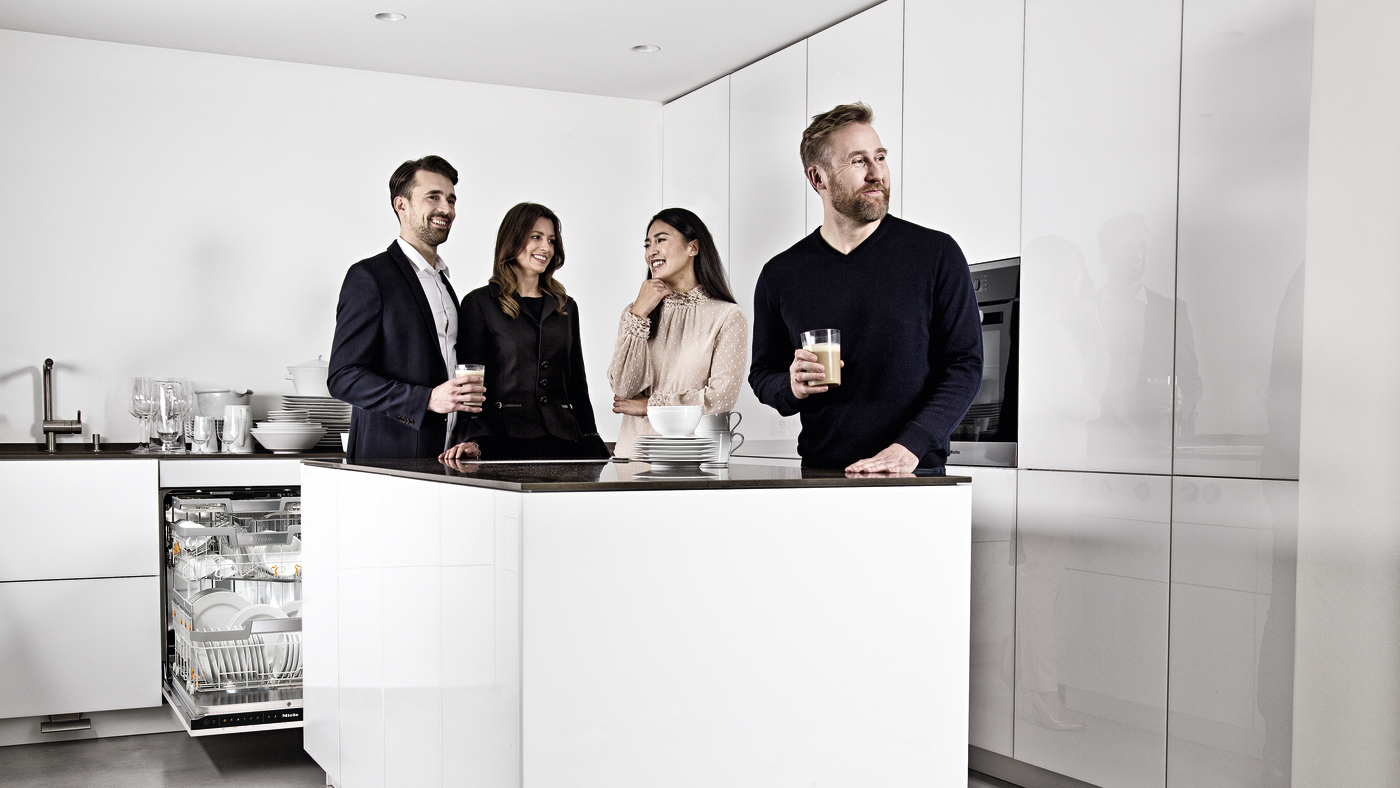 Four adults stand around a white kitchen island chatting, two holding drinks, with an open dishwasher showing clean dishes in the background.