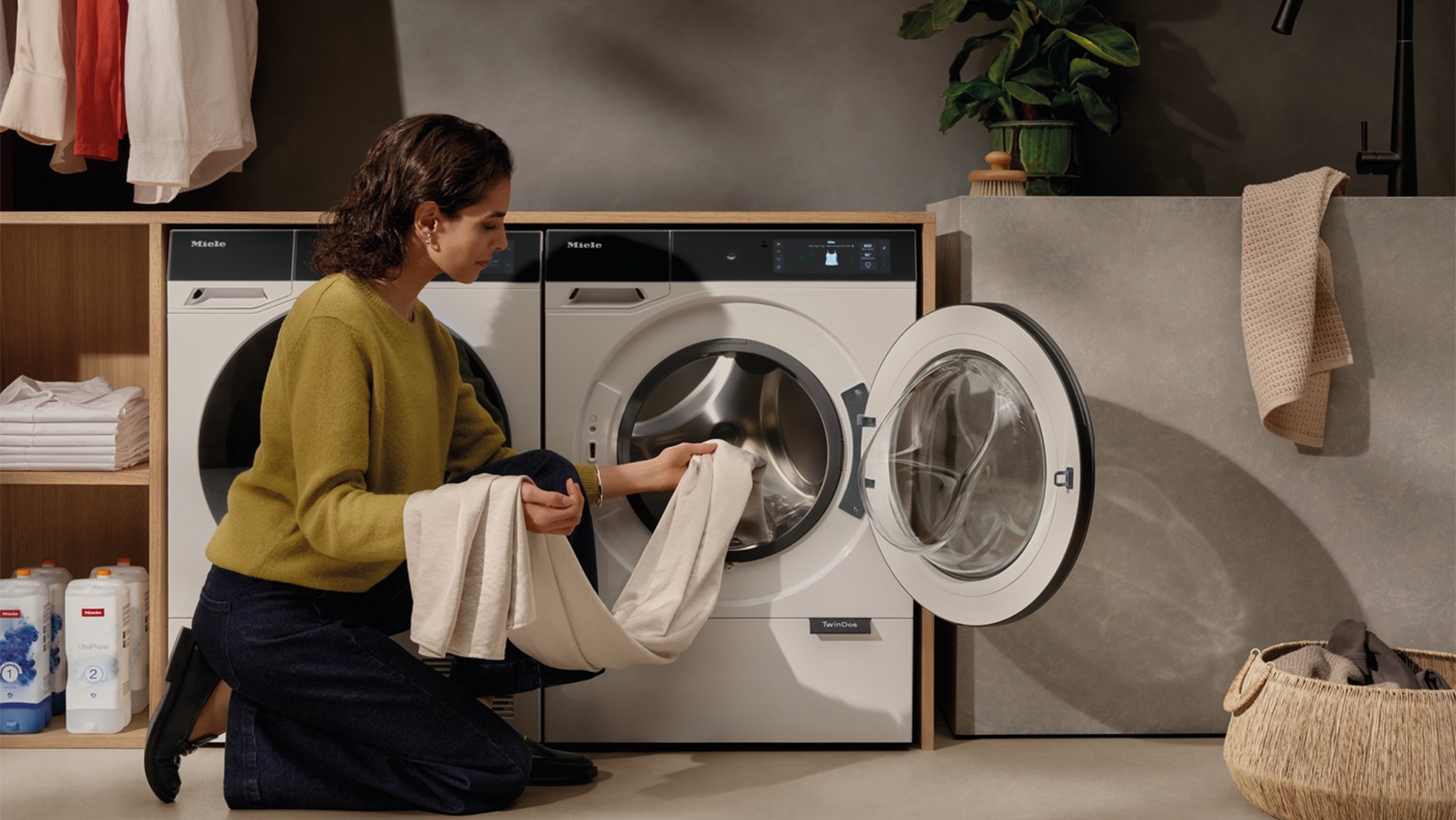 Woman kneels by an open front-loading washing machine, loading white laundry.