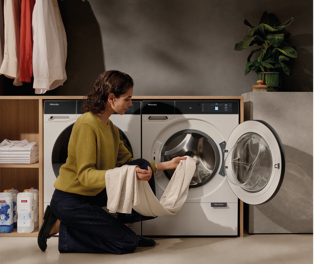 Woman kneeling and loading beige laundry into a front-loading Miele washing machine in a modern laundry room with shelves and a plant nearby.