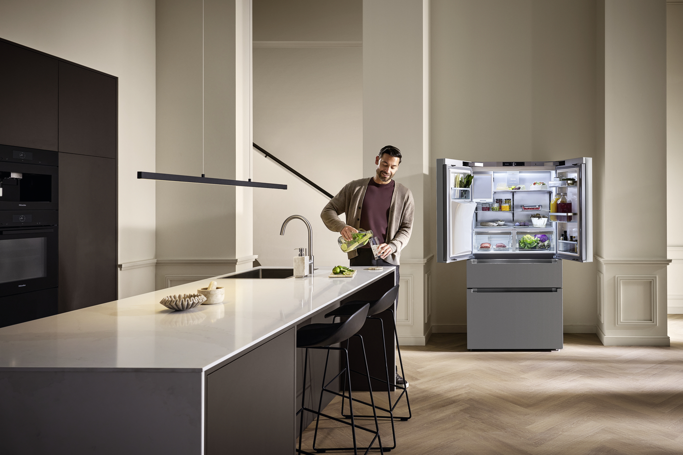 Man in a beige cardigan pours green juice into a blender at a sleek white kitchen island, with an open refrigerator showing produce nearby.