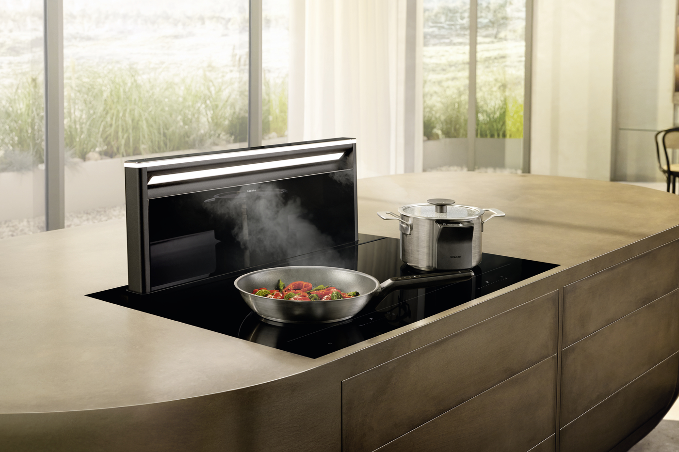 Induction cooktop built into a curved kitchen island with a skillet of peppers and greens sizzling on the surface, and a stainless pot nearby.