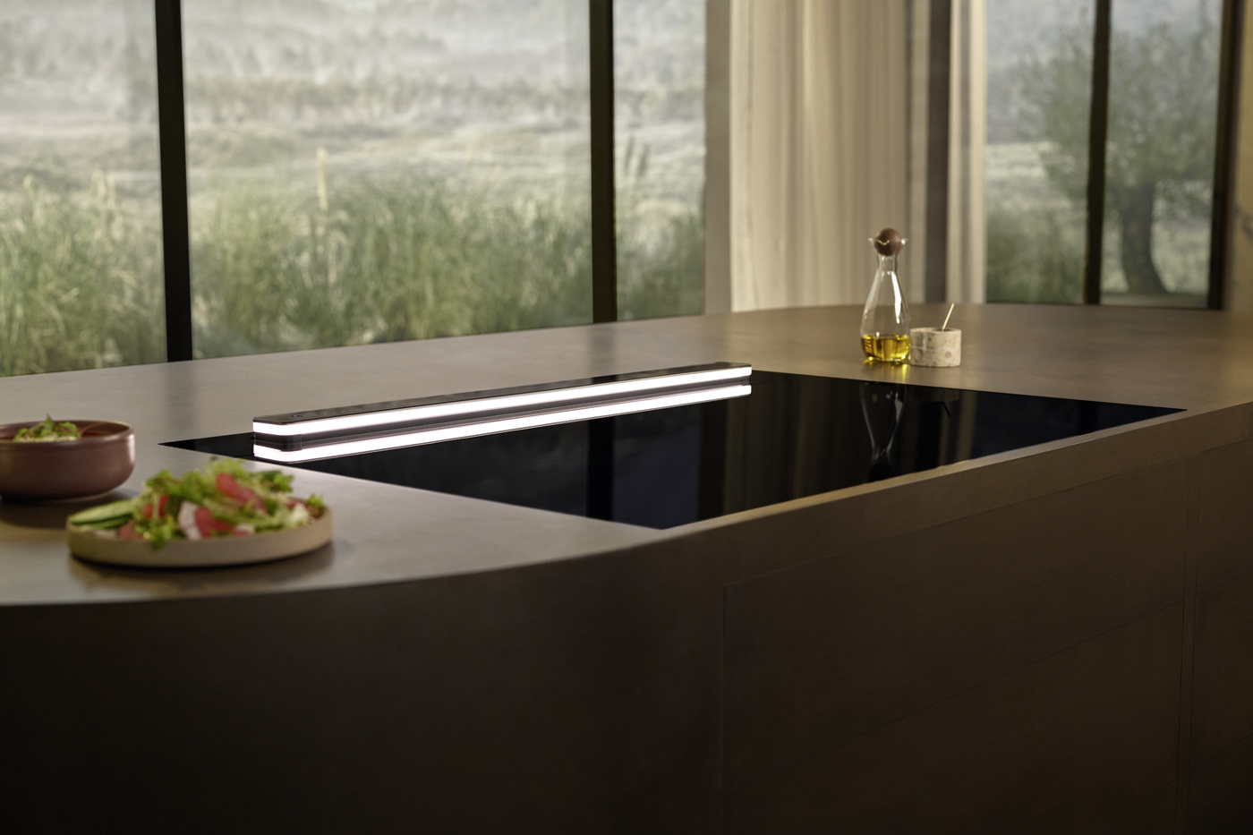 Modern kitchen island with a flush black induction hob, salads on the left and an oil bottle on the right near a window.