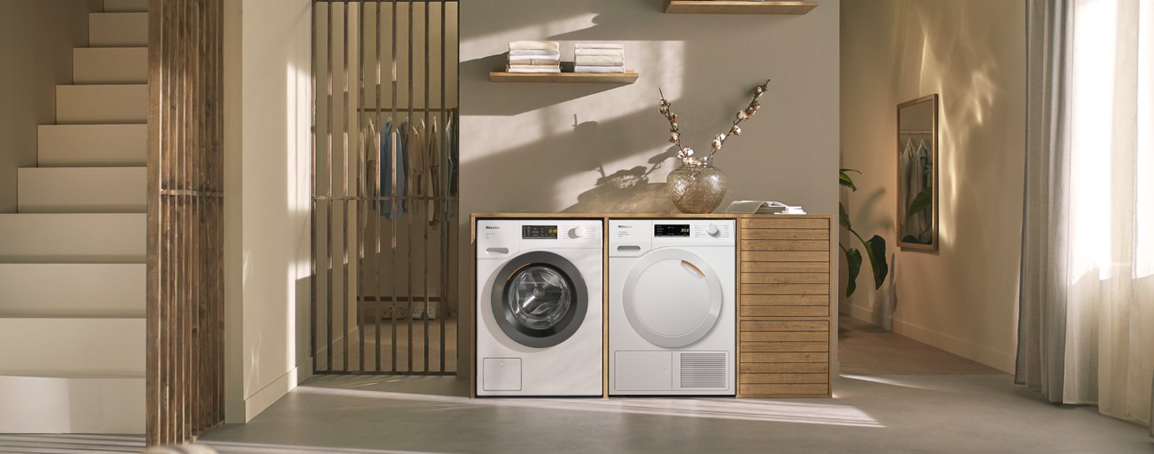 Modern laundry room with a front-loading washer and dryer beside a wooden cabinet, sunlit and minimalist.