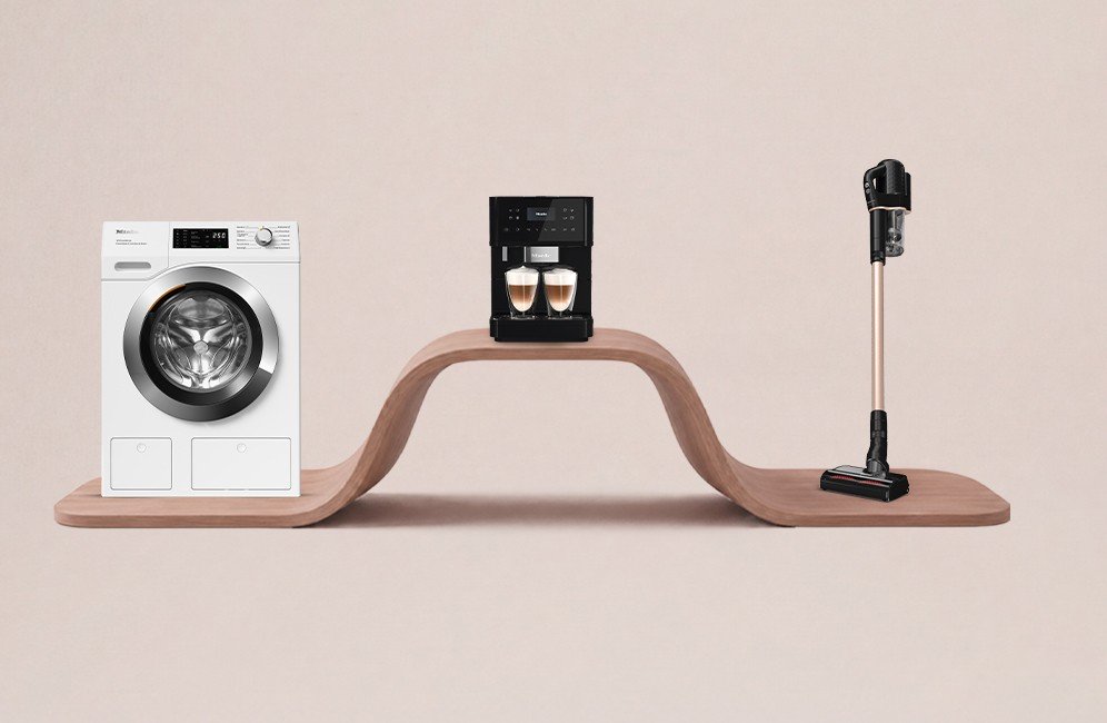 Wavy wooden shelf displaying a washing machine left, a coffee machine center, and a cordless vacuum on the right.
