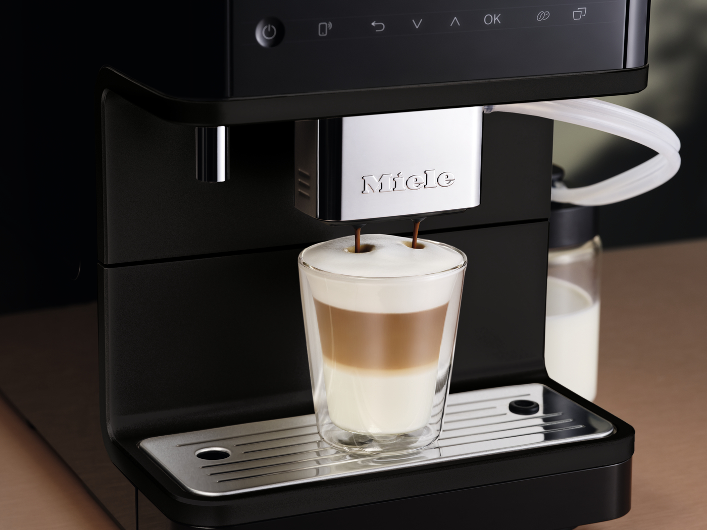 Miele espresso machine brewing a layered latte into a double‑walled glass cup on a metal tray with milk froth on top.