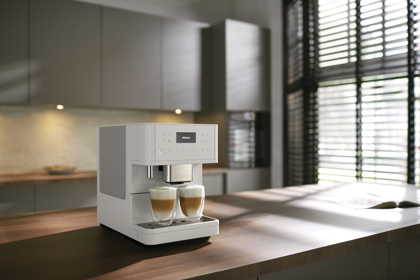 White Miele espresso machine on a wooden kitchen island brewing two cappuccinos with crema, in a modern sunny kitchen.