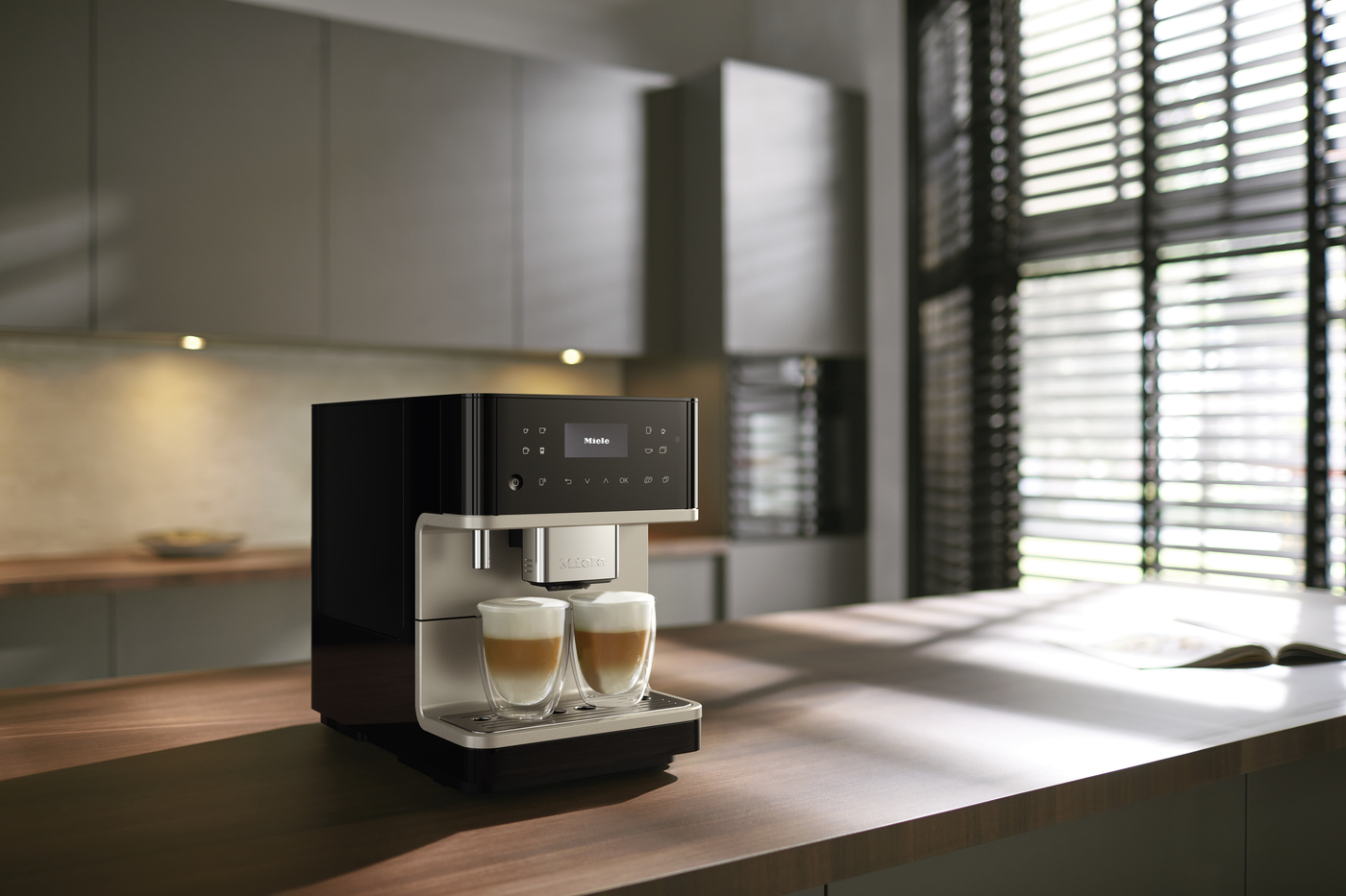 Miele espresso machine brewing two cappuccinos on a wooden kitchen island with sunlight streaming through blinds.