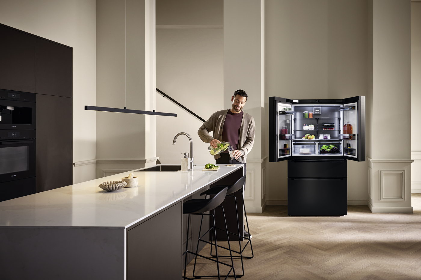 Man in a cardigan prepares vegetables at a sleek white kitchen island, with an open, well-stocked refrigerator in the background.