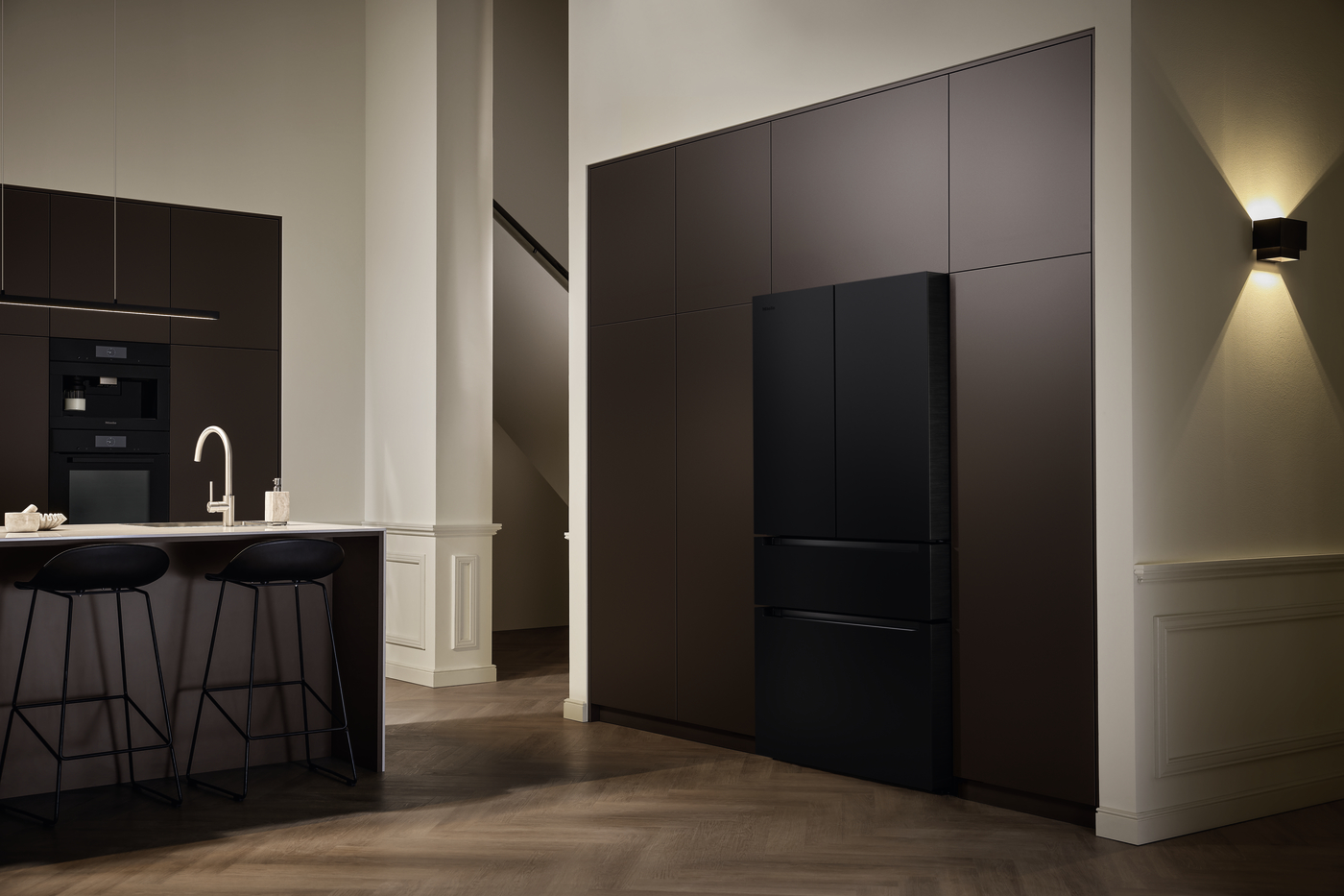 Modern kitchen with dark brown cabinetry, island sink, curved faucet, and two black bar stools at a white countertop island.