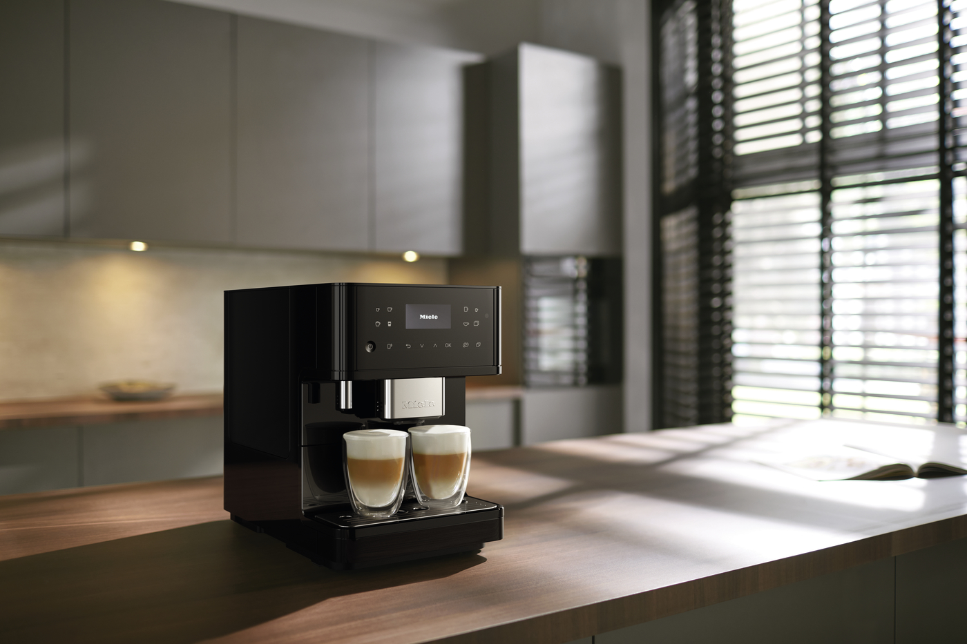 Black espresso machine on a wooden kitchen island brewing two cups of latte macchiato, with warm pendant lighting and blinds in the background.