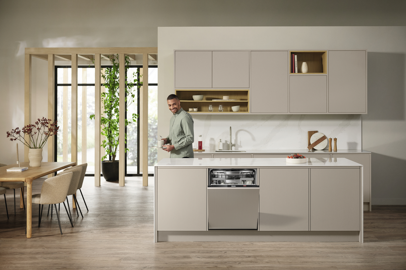 Man standing at a large light-gray kitchen island with a built-in dishwasher mid-open, in a modern open-plan kitchen and dining area.