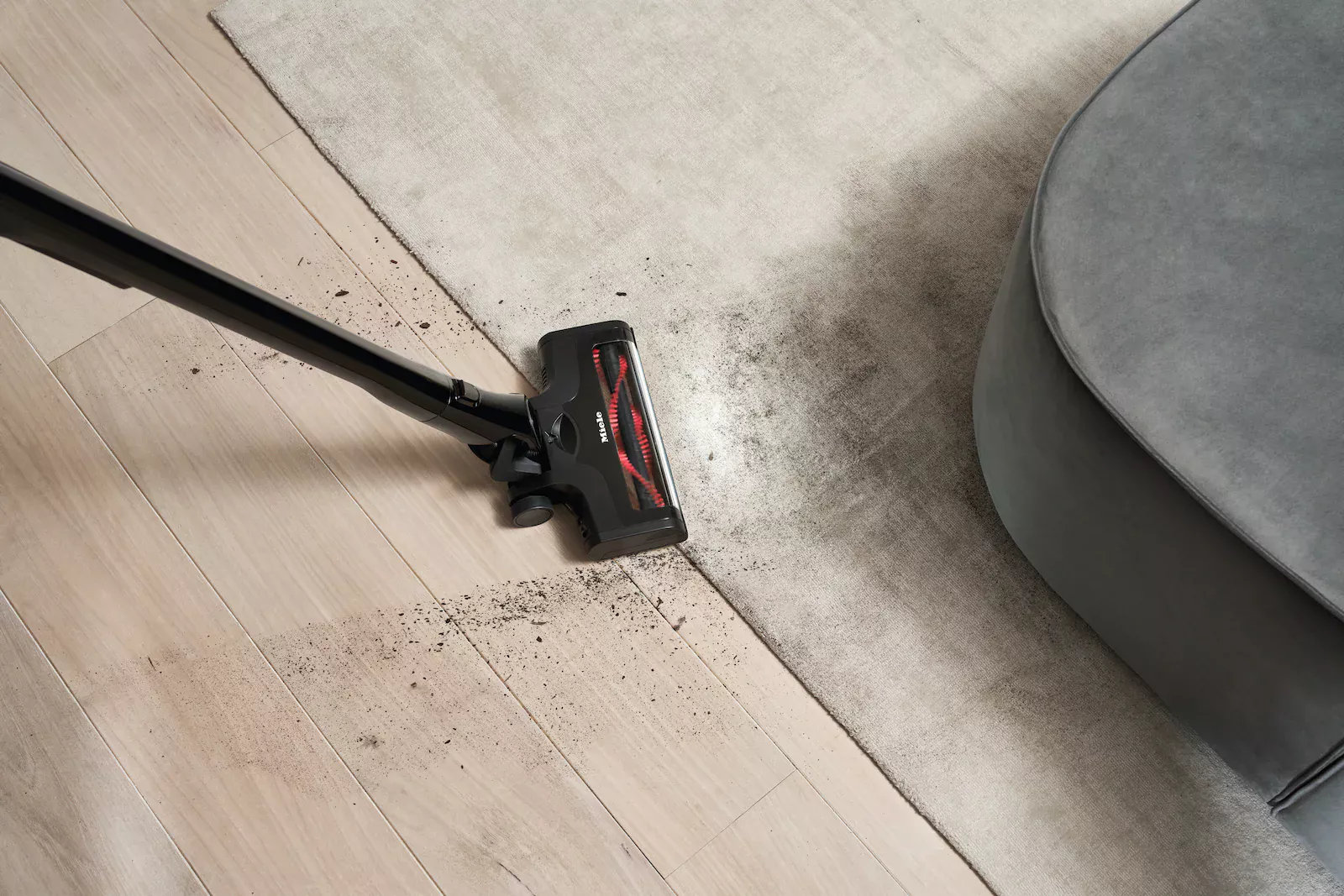 Cordless vacuum head cleaning dirt from a light wood floor and edge of a gray rug with a gray sofa nearby, showing dust trails left behind.