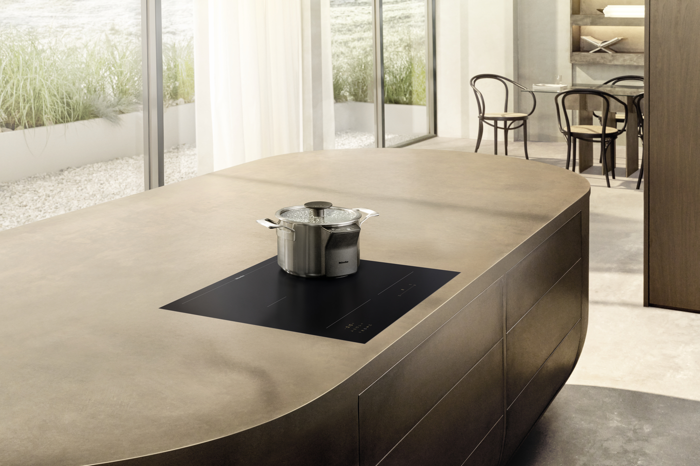 Copper-toned kitchen island with a black induction cooktop and a stainless steel pot on top, in a bright open dining area and garden view outside