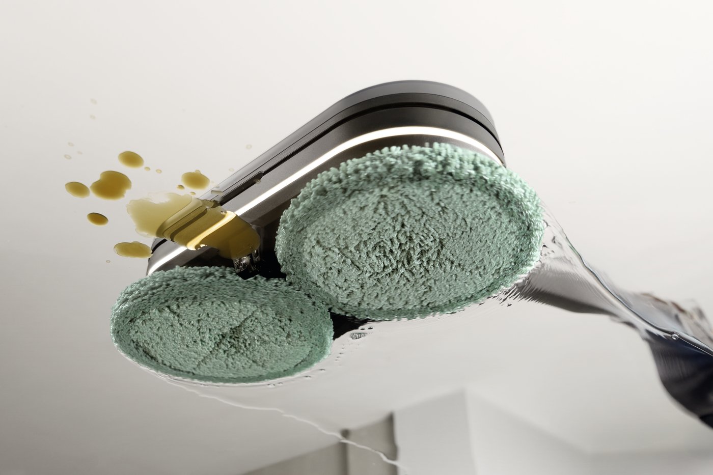 Close-up of a cleaning head with two green sponge pads spraying yellow liquid on a white ceiling, highlighting a cleaning action.