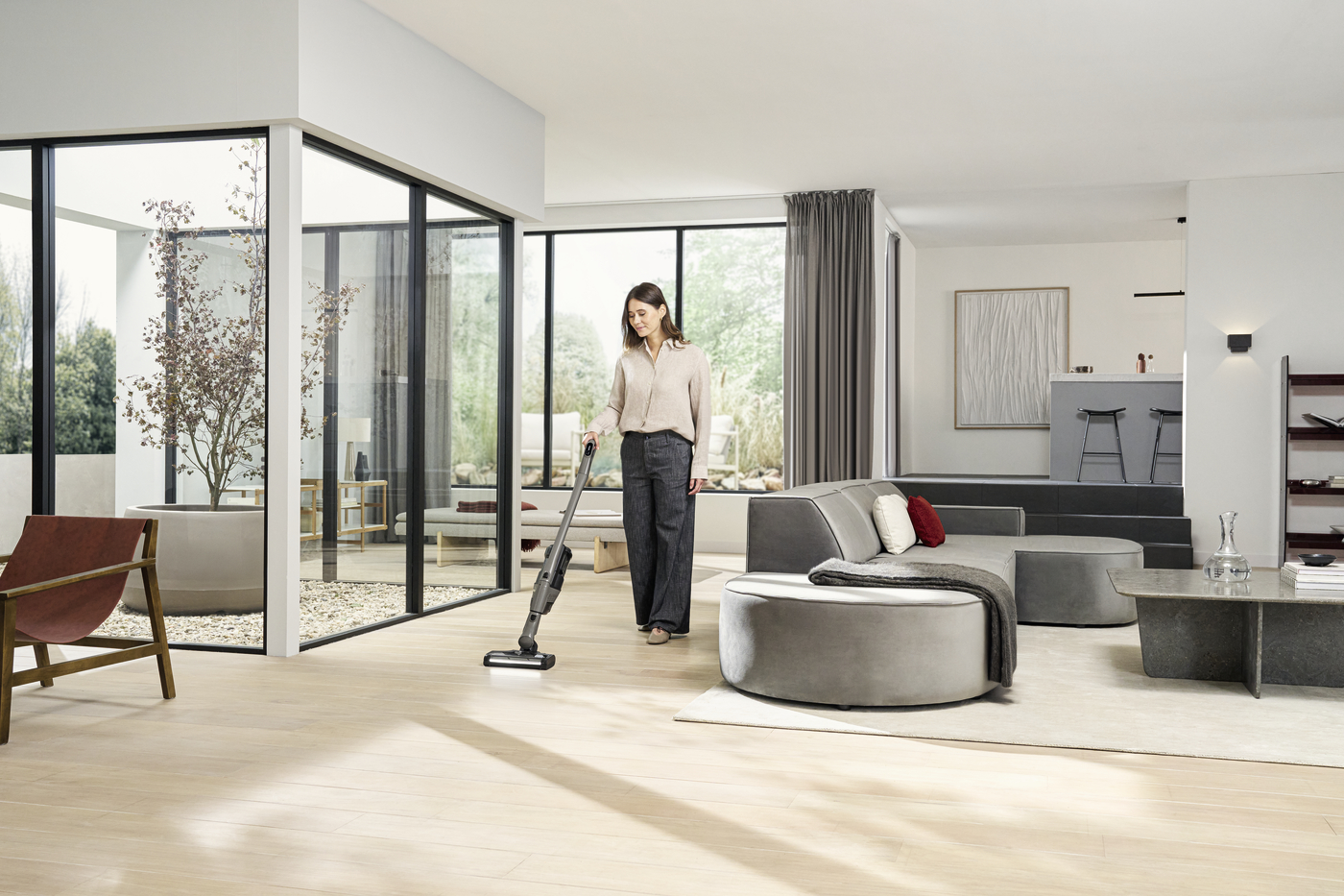 Woman vacuuming a modern open-plan living room with large glass walls and natural light.