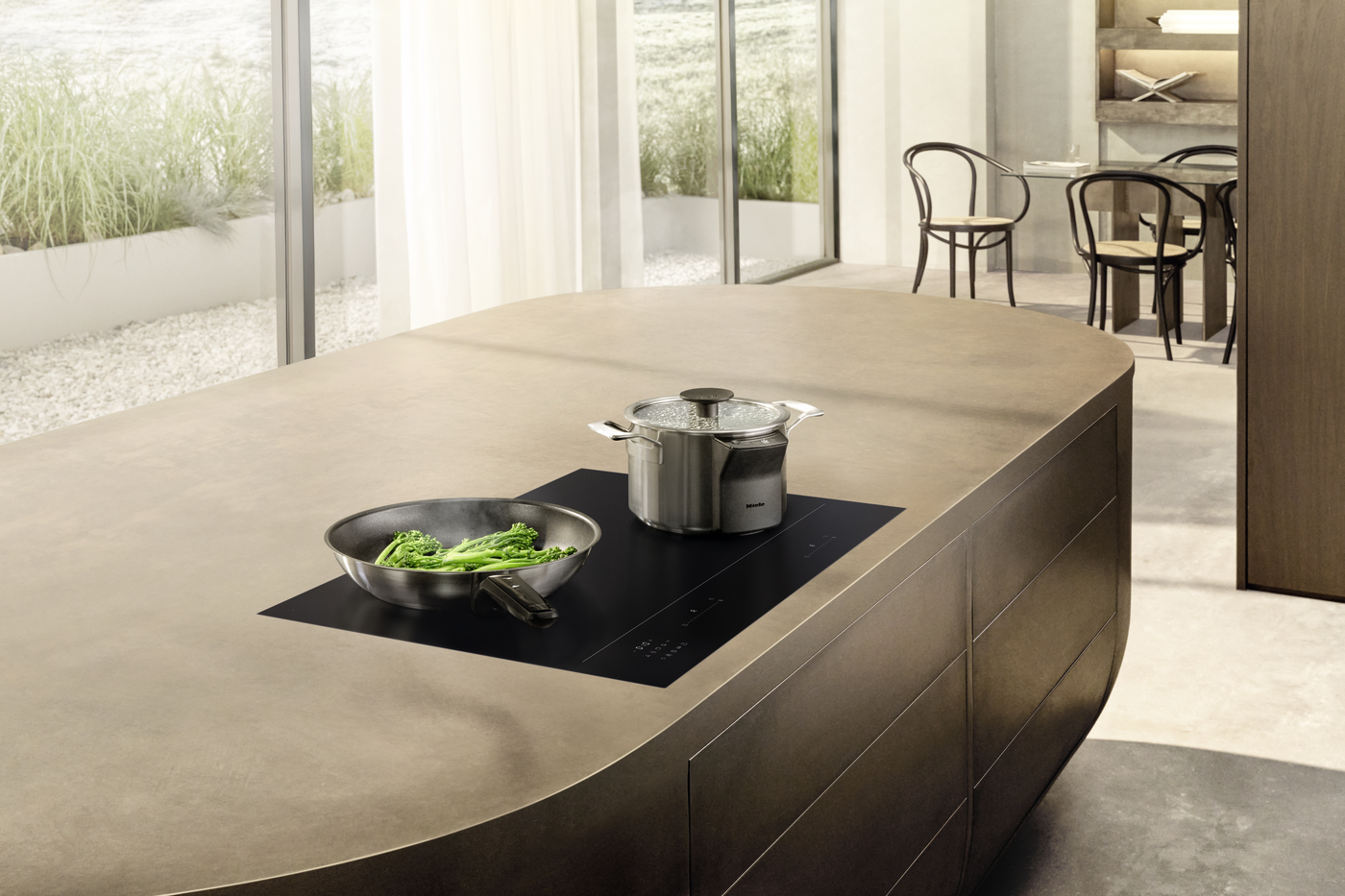 Modern kitchen island with a black induction cooktop; a stainless steel pot and a pan of broccoli sit on the surface, sunlit by large windows.