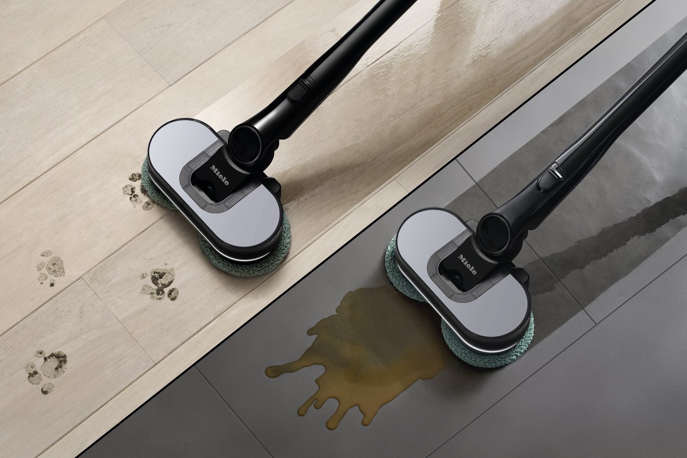Two Miele floor scrubbers with green pads actively clean a brown spill on a dark floor surface nearby a wooden floor edge