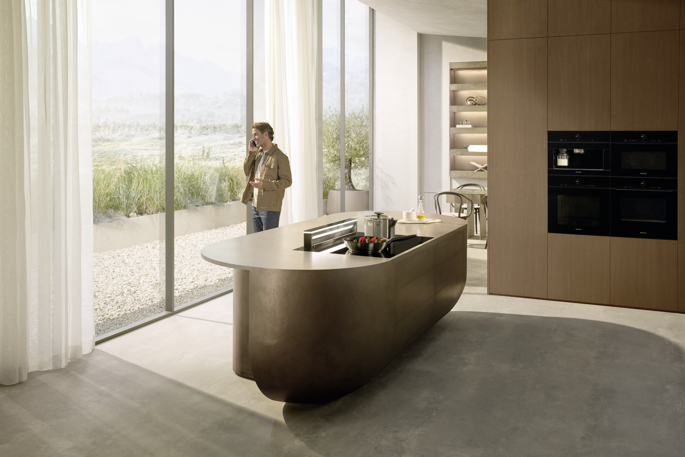 Man on the phone standing at a curved bronze kitchen island in a bright modern kitchen with floor-to-ceiling windows.