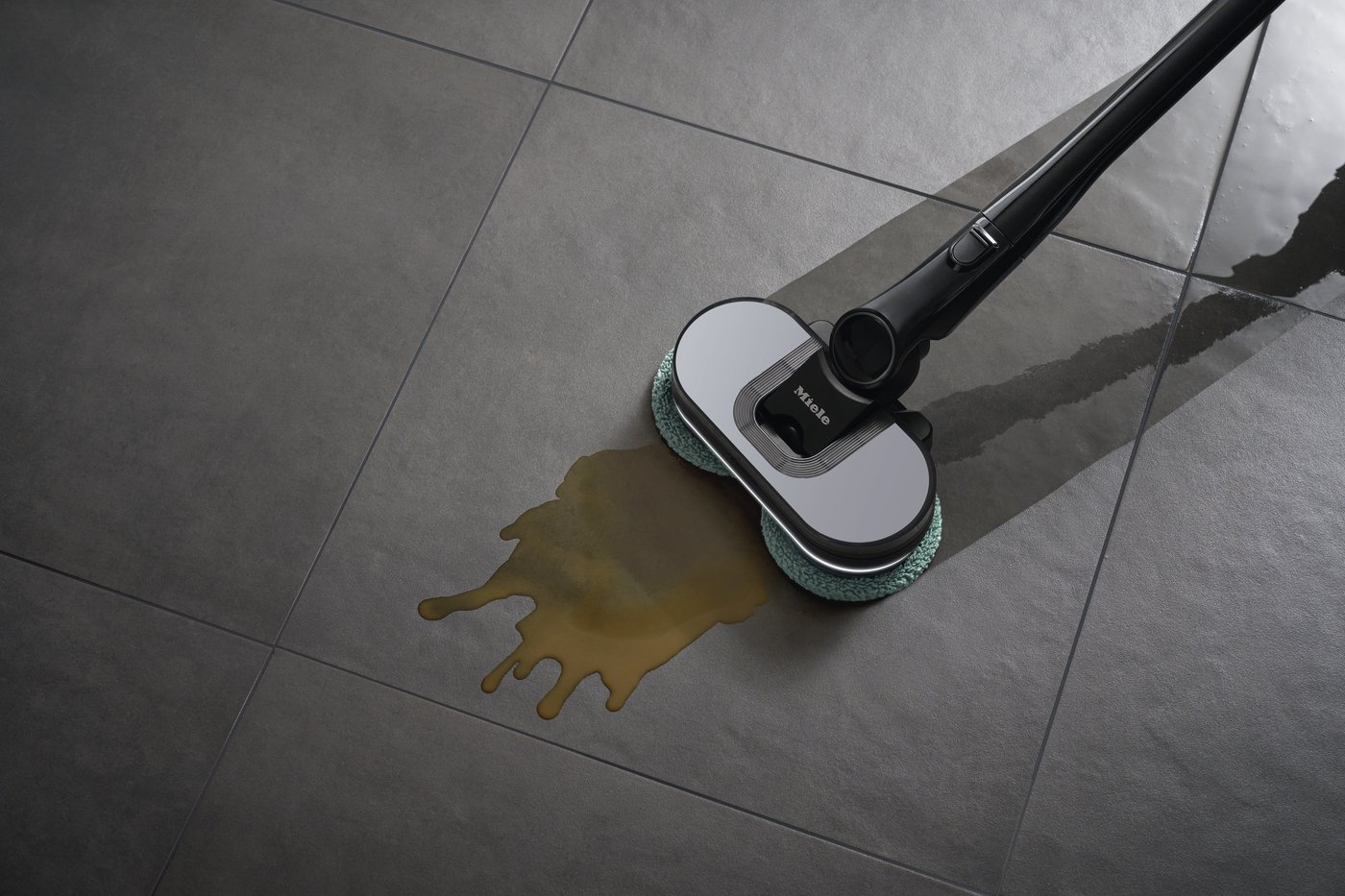 Steam mop with green pad cleaning a brown liquid spill on gray tile floor