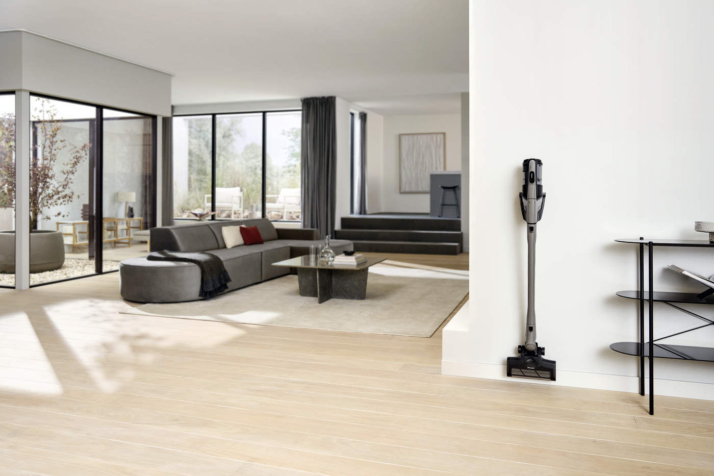 Open, airy living room with a gray sectional, glass coffee table, and large windows; a vacuum cleaner stands against a white pillar on the right.
