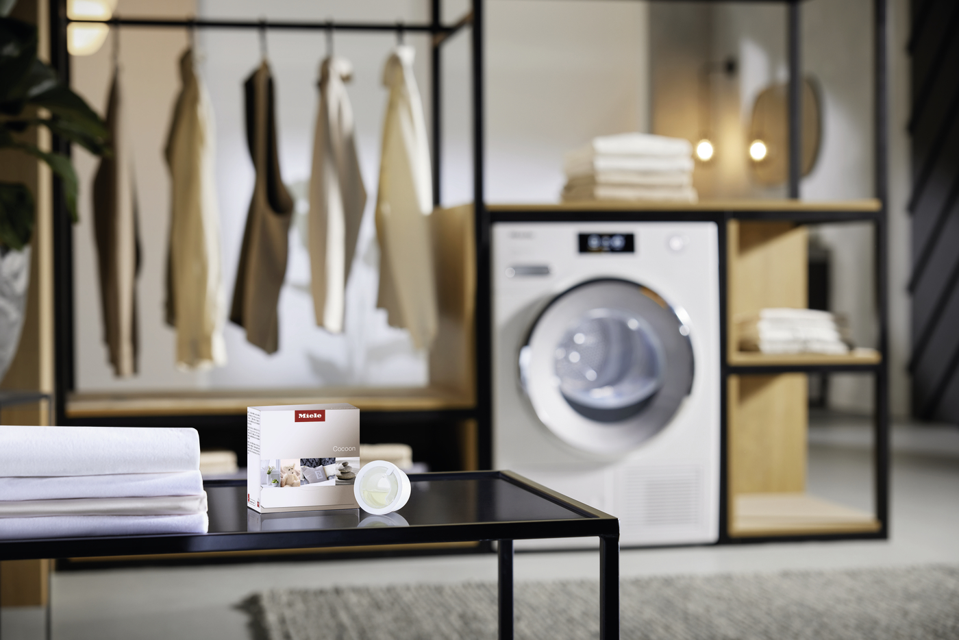 Showroom with a front-loading washing machine in a black-wood shelving unit; foreground features folded towels and a Miele Cocoon box on a glass table, with beige clothing hanging in the background.
