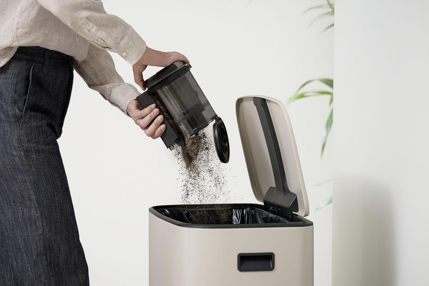 Person pours a scoop of coffee grounds into a beige trash can with the lid open and a black liner inside.