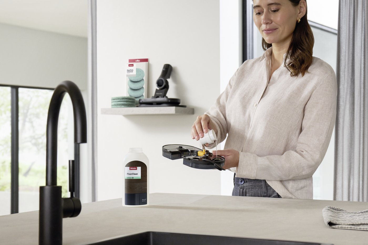 Woman in a beige sweater pours liquid into a cleaning tool on a kitchen island; a FloorClean bottle sits nearby on the counter.