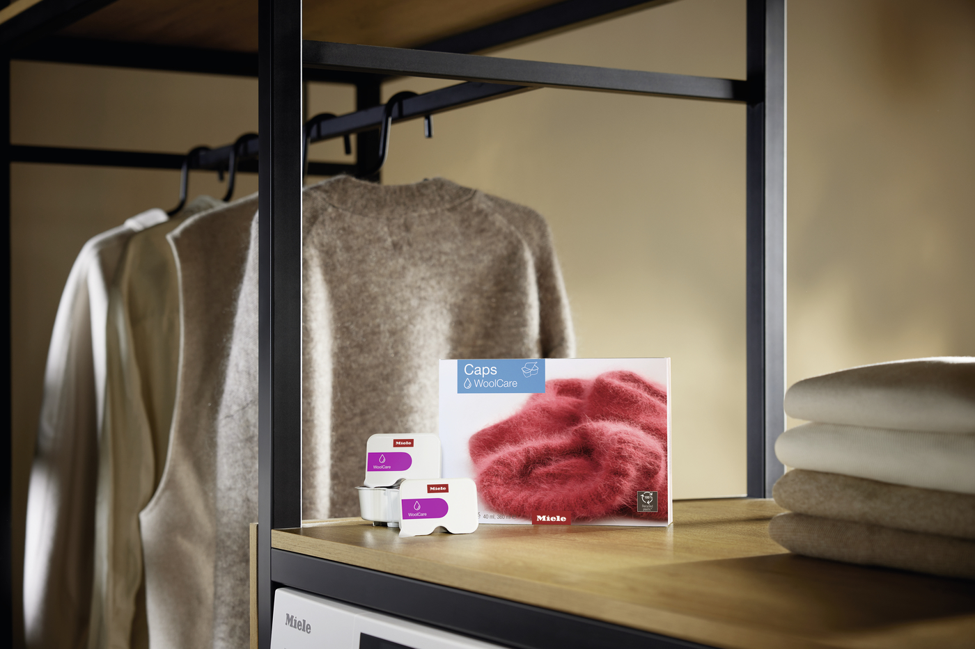 Wooden shelf with a brown sweater on a hanging rack, folded towels nearby, and a WoolCare Caps box featuring red wool with small care bottles in front.