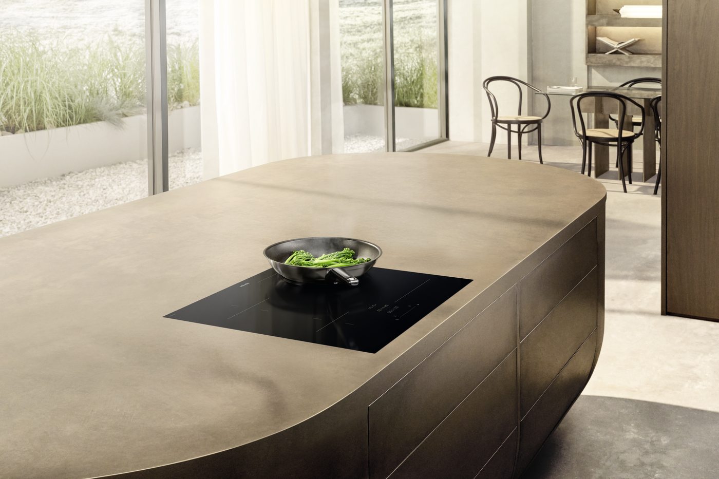 Modern kitchen island with a sleek black induction cooktop and a pan of green vegetables, in a bright open-plan room.
