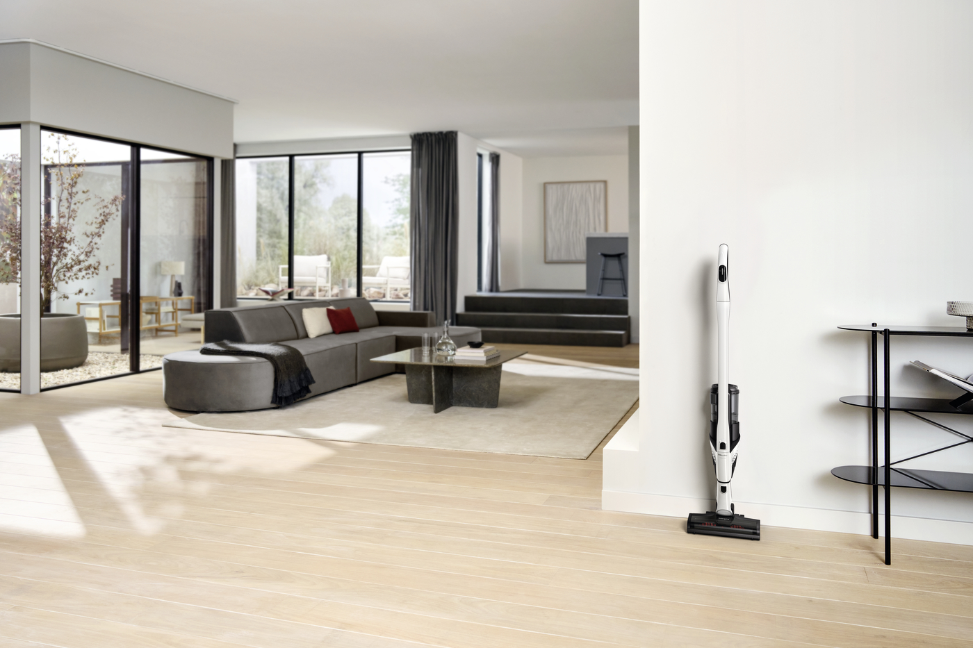 Bright open living room with large floor-to-ceiling windows, gray sectional sofa, and a vacuum standing near a white wall on the right side in a modern home.